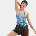 Woman wearing a blue patterned top and brown shorts with a black Coach handbag on a white background
